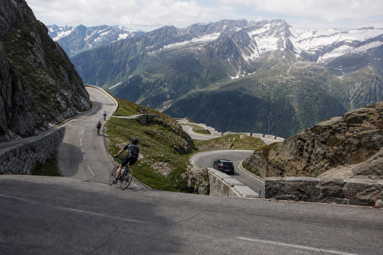 Cyclistes et voiture dans les lacets d'un col alpin, face à des sommets encore enneigés au début de l'été